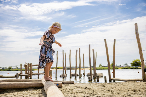 Una niña camina descalza sobre un tronco en la playa de un parque vacacional con glamping junto al lago.