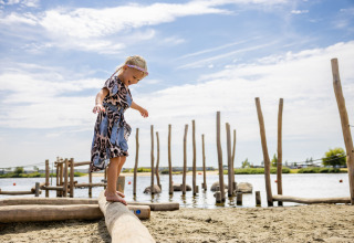 A child balances barefoot on a wooden beam at the lakeside beach of a glamping holiday park.