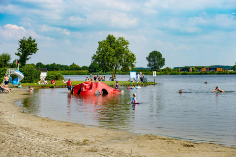 Niños y adultos disfrutan en un lago de un parque vacacional con glamping y divertidas zonas recreativas.