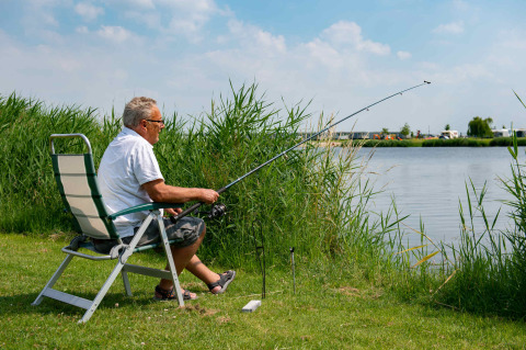 Older man fishing calmly from a chair by a lakeside in a holiday park with glamping accommodations.