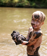 Niño alegre cubierto de barro juega junto al río en un parque vacacional con alojamiento glamping.