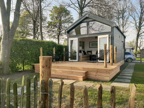 Kleines Lodge namens Tiny House mit Holzterrasse und Gartenstühlen, umgeben von Bäumen und Hecken.