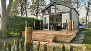 Kleines Lodge namens Tiny House mit Holzterrasse und Gartenstühlen, umgeben von Bäumen und Hecken.