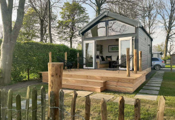 Kleines Lodge namens Tiny House mit Holzterrasse und Gartenstühlen, umgeben von Bäumen und Hecken.