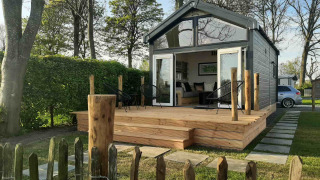 Tiny house lodge with wooden deck and outdoor chairs, surrounded by green trees and a hedge in the garden.