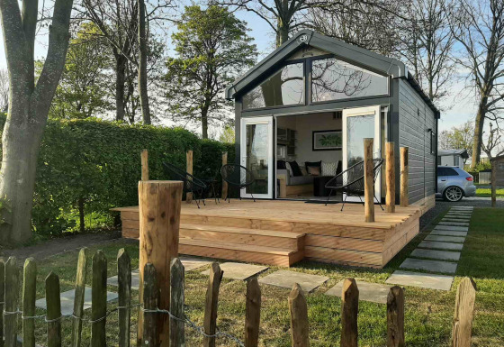 Casa pequeña llamada Tiny House con terraza de madera y sillas, rodeada de césped y árboles verdes.
