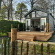Lille træhytte kaldet Tiny House med terrasse og havesiddepladser, omgivet af grønne træer og hæk.