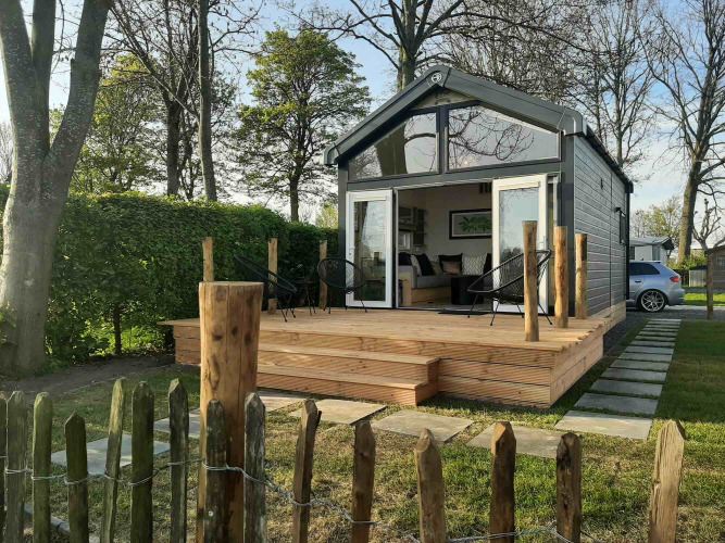 Casa pequeña llamada Tiny House con terraza de madera y sillas, rodeada de césped y árboles verdes.