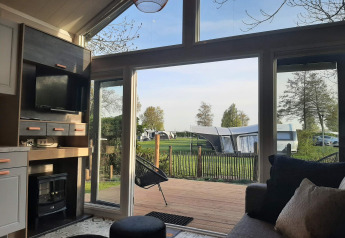 Udsigt fra en hyggelig Tiny house lodge med store vinduer ud til campingplads og grønne omgivelser.