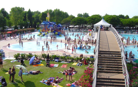Busy outdoor pool with slides, sunbathers on grass, and a footbridge at Camping Village Marina di Venezia, Italy.