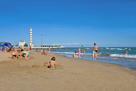 Scena di spiaggia al Camping Village Marina di Venezia, Veneto, Italia, con famiglie e un faro sullo sfondo.