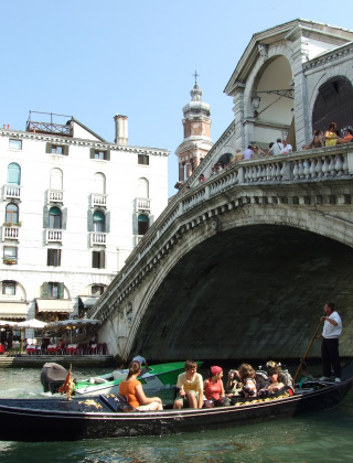 Una góndola navega bajo el Puente de Rialto en Venecia, Italia, con turistas en la embarcación y el puente.