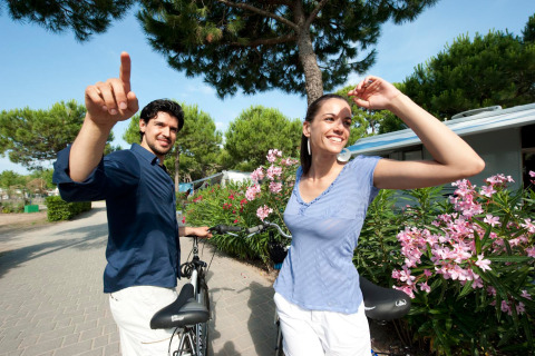 Una pareja sonriente con bicicletas disfruta de un día soleado en Camping Village Cavallino, rodeados de flores.
