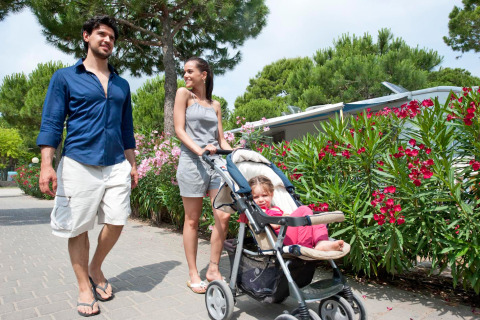 Familia paseando por Camping Village Cavallino en Veneto, Italia, con niño en cochecito y flores alrededor.