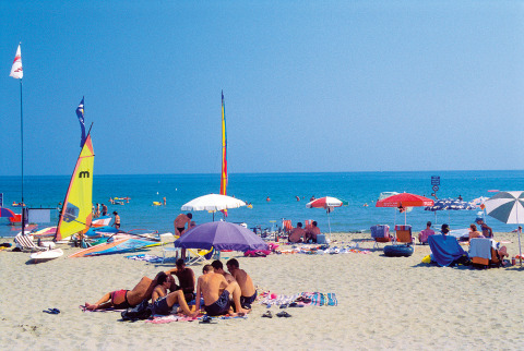 Personas descansando en la playa de arena de Camping Village Cavallino, Véneto, Italia, con sombrillas y veleros.