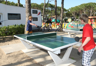 Due ragazzi giocano a ping pong all'aperto al Camping Village Cavallino, con camper e parco giochi colorato sullo sfondo.