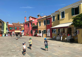 Børn leger på en åben plads omgivet af farverige huse i solskin, Camping Village Cavallino, Veneto, Italien.