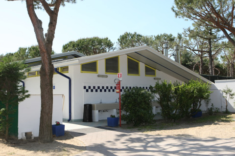 Edificio de baños exteriores en Camping Village Cavallino, Veneto, Italia, rodeado de árboles y vegetación.