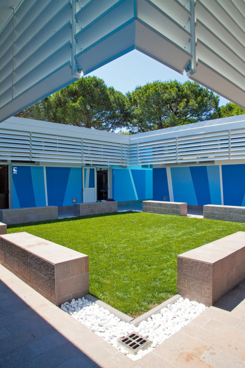 Patio interior con césped, bancos de piedra y paredes azules en Camping Village Cavallino, Véneto, Italia.