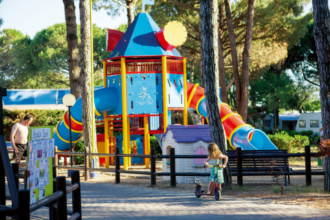 Niños juegan en un colorido parque infantil en Camping Village Cavallino, rodeados de altos árboles verdes.