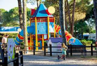 Children enjoy a colorful playground at Camping Village Cavallino holiday park, surrounded by tall trees.