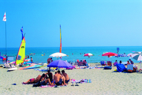 Día soleado en la playa del Camping Village Cavallino, Veneto, Italia, con sombrillas, bañistas y veleros.