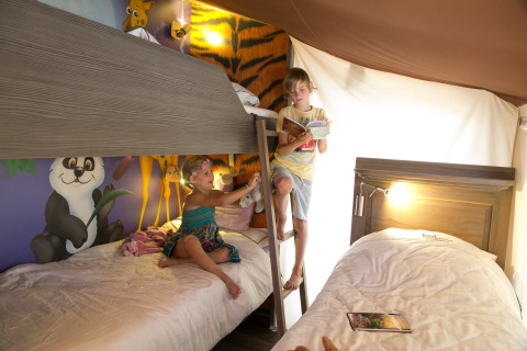 Niños leyendo y jugando en una colorida cabaña temática de jungla con literas en Camping Village Cavallino, Italia.