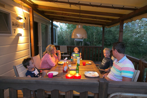 Una familia cena junta en una terraza acogedora con techo de una cabaña, iluminada por luces cálidas.