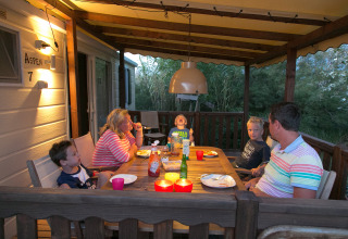 Una familia cena junta en una terraza acogedora con techo de una cabaña, iluminada por luces cálidas.