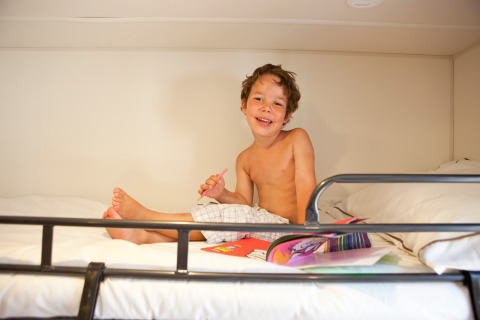 Jongen zit lachend te tekenen op een bed in SunLodge Redwood, Camping Village Cavallino, Italië.