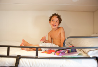 Garçon souriant assis sur un lit et dessinant à SunLodge Redwood, Camping Village Cavallino, Italie.