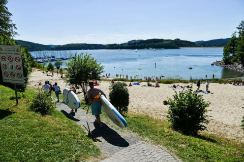 Parque de vacaciones con glamping, personas llevando tablas de paddle hacia la playa junto a un lago soleado.