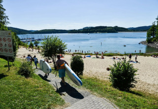 Parque de vacaciones con glamping, personas llevando tablas de paddle hacia la playa junto a un lago soleado.