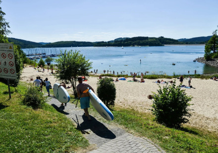 Vakantiepark met glamping, mensen dragen paddleboards richting het zandstrand aan een zonnig meer.