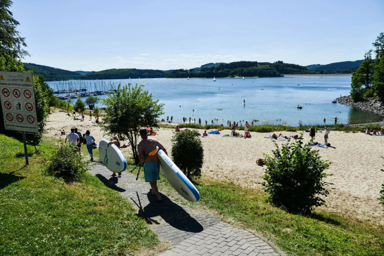 Ferienpark mit Glamping, Menschen tragen Paddleboards zum Strand an einem sonnigen Tag am See.
