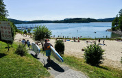 Holiday park with glamping, people carrying paddleboards down to a sandy beach by a lake in summer sun.