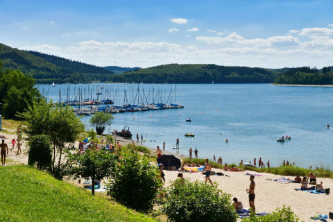 Playa soleada junto al lago con bañistas, veleros y gente descansando, ideal para vacaciones glamping.