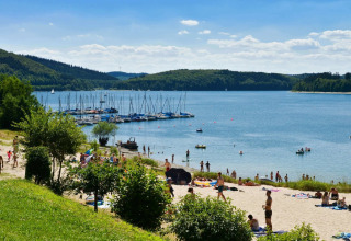 Solrig strand ved sø med svømmere, solbadende og sejlbåde, perfekt til ferie og glamping i naturen.