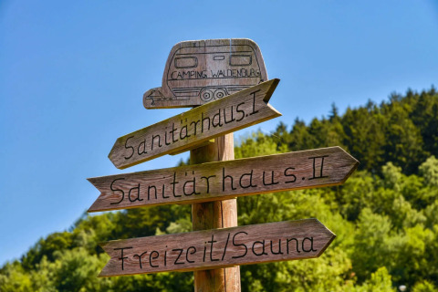 Panneau en bois au camping indiquant les sanitaires et sauna, sur fond de forêt et ciel bleu.
