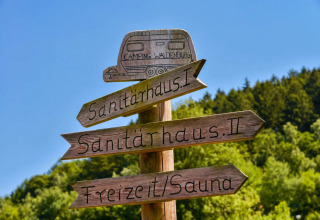 Panneau en bois au camping indiquant les sanitaires et sauna, sur fond de forêt et ciel bleu.