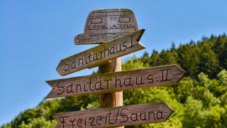 Letrero de madera en un camping con direcciones a edificios sanitarios y sauna, rodeado de vegetación.