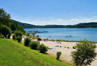 Playa en parque vacacional con glamping, colinas verdes, bañistas y veleros en un lago tranquilo de verano.