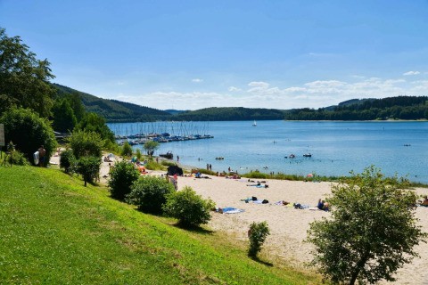 Strand ved feriepark med glamping, grønne bakker, badende gæster og sejlbåde på en rolig sø om sommeren.