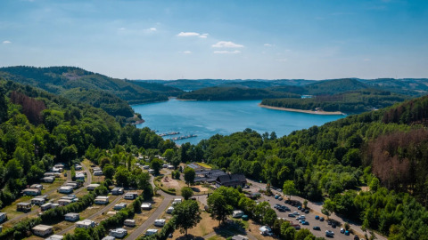 Vakantiepark met glamping aan een meer, omgeven door bossen en heuvels, luchtfoto op een zonnige dag.