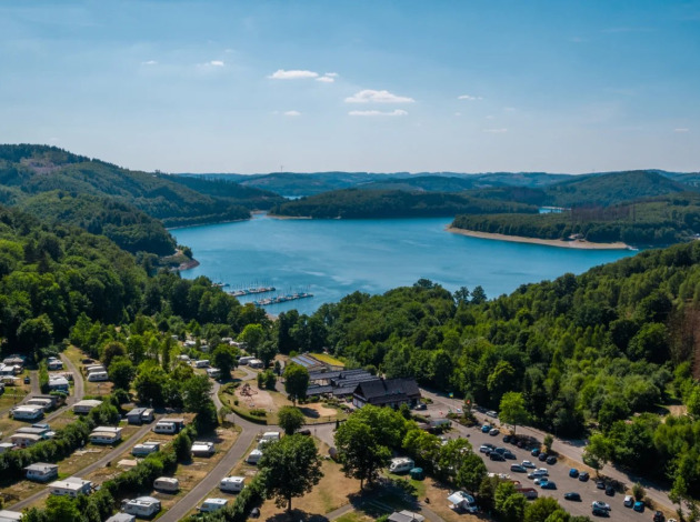Vakantiepark met glamping aan een meer, omgeven door bossen en heuvels, luchtfoto op een zonnige dag.