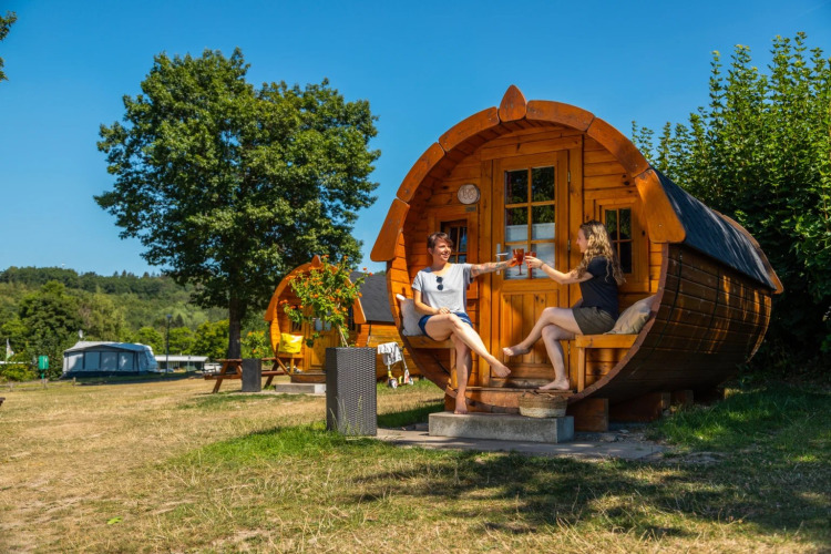 Zwei Frauen stoßen mit Getränken vor einer Holz-Glampinghütte im Ferienpark an einem Sommertag an.
