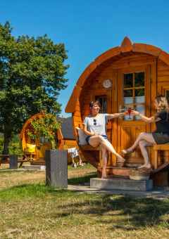 Dos mujeres brindan con bebidas frente a una cabaña glamping de madera en un parque vacacional soleado.
