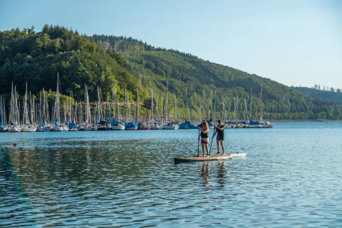 Two people stand paddleboarding on a lake in front of many sailboats and green hills at a holiday park.