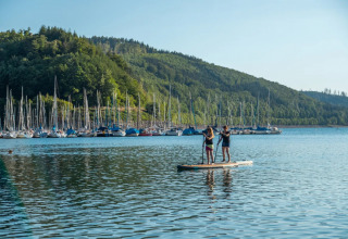 Due persone fanno paddleboard su un lago davanti a molte barche a vela e colline verdi in un villaggio turistico.