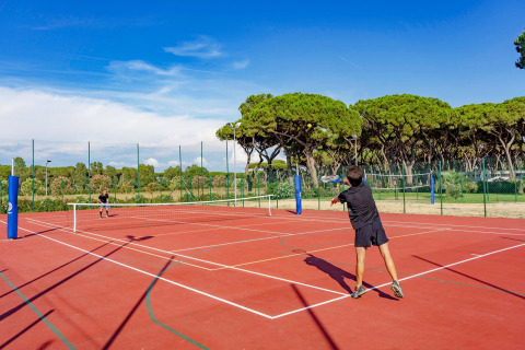 Two people playing tennis on an outdoor court surrounded by trees at Camping Village Roma Capitol in Tuscany, Italy.